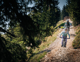 Gruppe von Mountainbiker:innen fährt einen steinigen Trail im Bikepark hinab, eingerahmt von dichten Bäumen und alpiner Landschaft. | © Yvonne Hörl