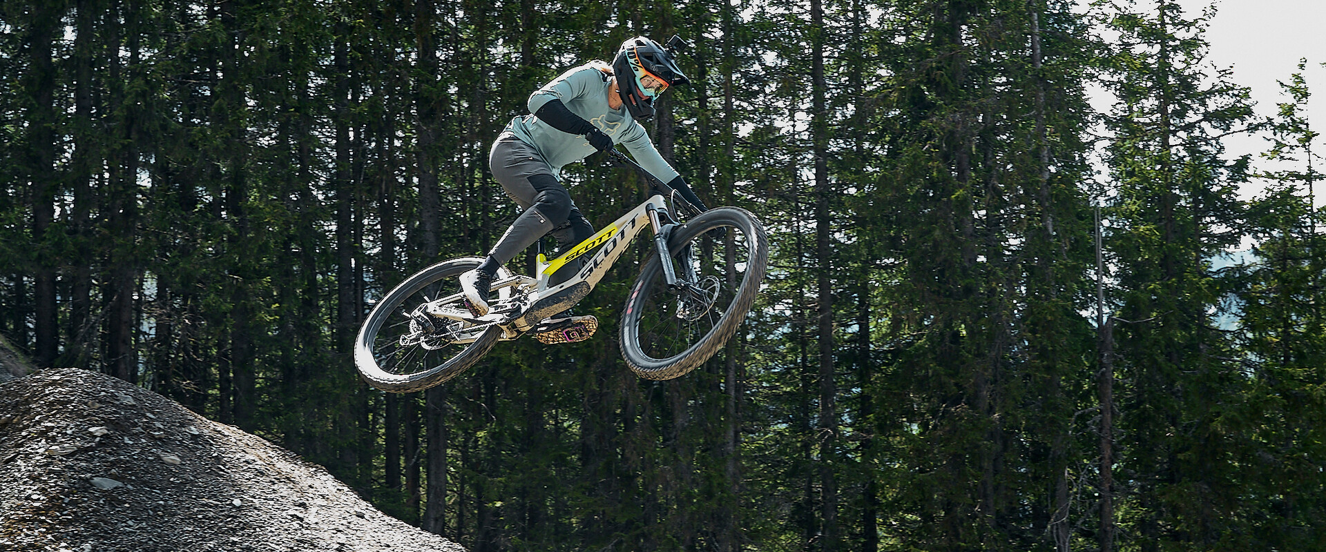 Mountainbikerin mit Vollvisierhelm und Protektoren springt mit ihrem Downhill-Bike über einen Kicker im Bikepark, umgeben von dichtem Wald. | © Karin Pasterer