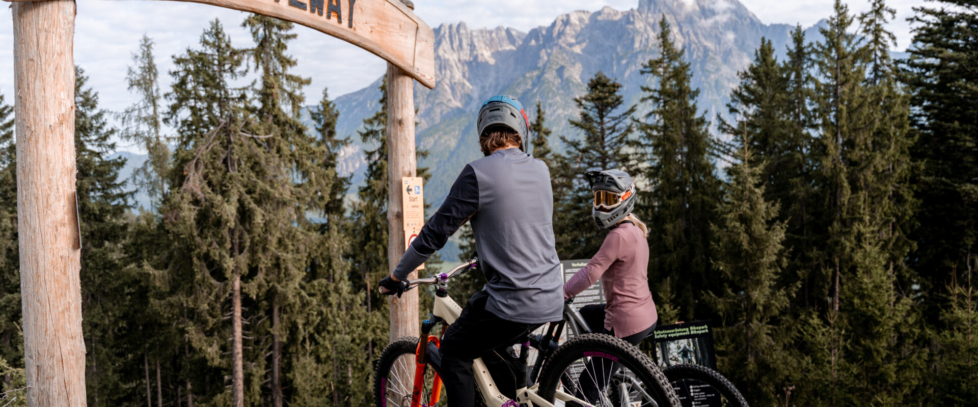 Zwei Mountainbiker:innen mit Fullface-Helmen stehen mit ihren Bikes unter dem hölzernen „Gateway“-Bogen am Start eines Trails im Bikepark Leogang mit Blick auf das Steinerne Meer. | © Yvonne Hörl