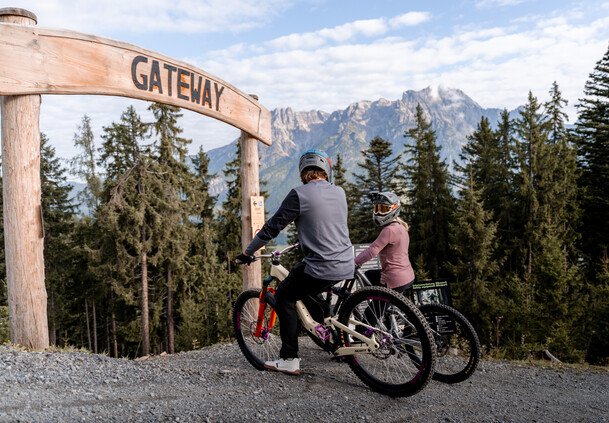 Zwei Mountainbiker:innen mit Fullface-Helmen stehen mit ihren Bikes unter dem hölzernen „Gateway“-Bogen am Start eines Trails im Bikepark Leogang mit Blick auf das Steinerne Meer. | © Yvonne Hörl