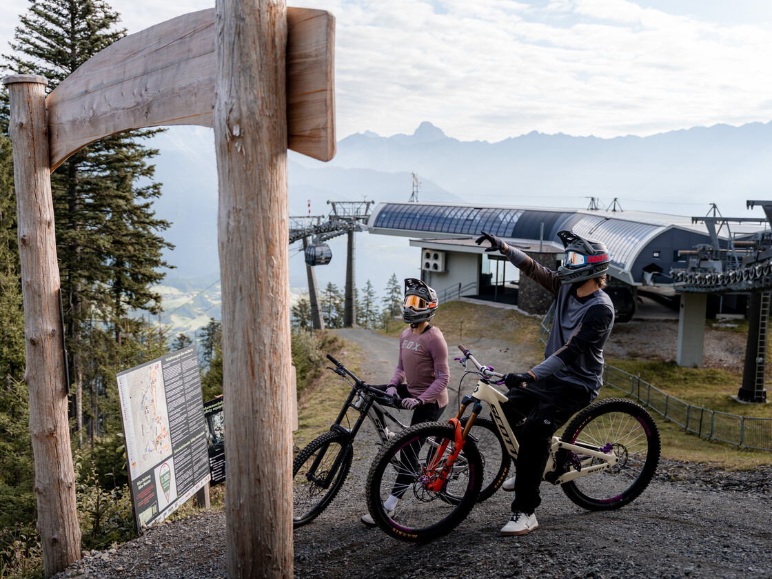 Zwei Biker:innen mit Helmen studieren die Trailmap unter dem hölzernen Gateway-Bogen im Bikepark Leogang, mit der Bergstation und Gondel im Hintergrund. | © Yvonne Hörl