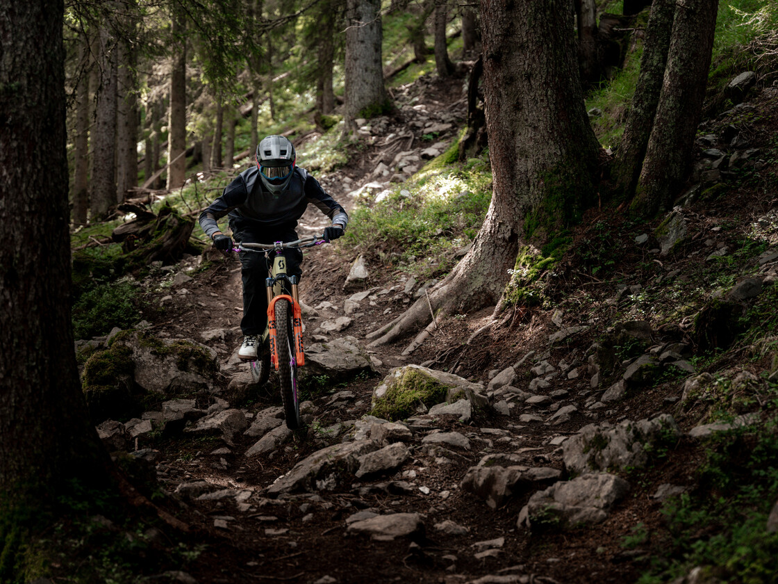 Ein Biker mit weißem Fullface-Helm fährt über steiniges, unebenes Terrain zwischen Wurzeln und Felsblöcken auf einem naturbelassenen Downhill-Trail.